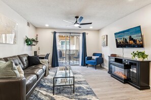 A living room with a leather sofa, a glass coffee table, a blue armchair, a fireplace, and a cityscape painting.