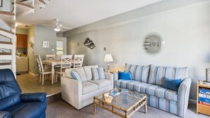 Una sala de estar con un sofá, una mesa de centro, una silla azul, un comedor con una mesa y sillas, y una escalera.