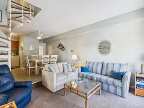 Ein Wohnzimmer mit einer Couch, einem Couchtisch, einem blauen Sessel, einem Essbereich mit Tisch und Stühlen sowie einer Treppe.