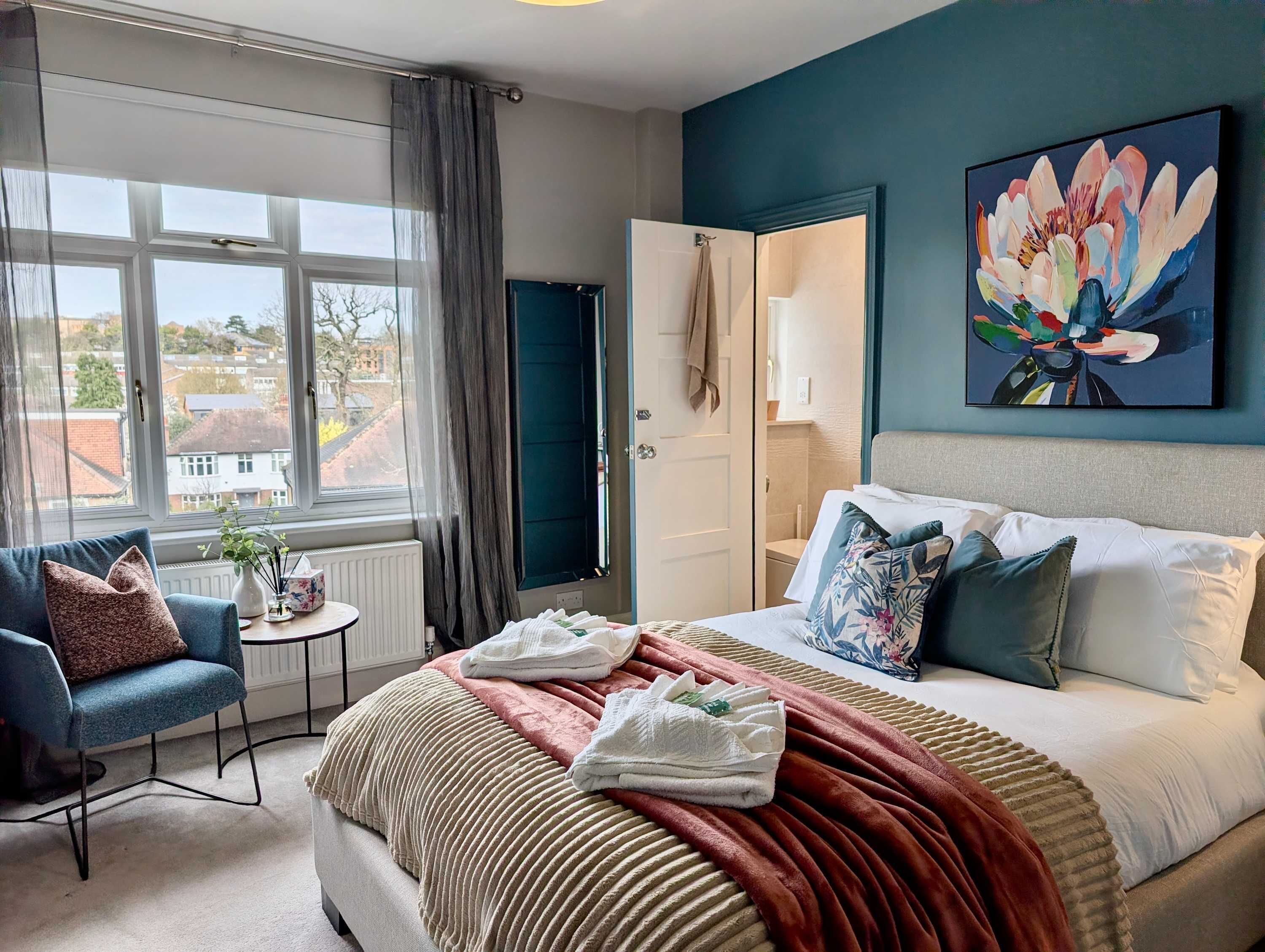 A hotel room with a bed, a chair, a small table, a window with curtains, and a painting on the wall.
