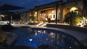 Une piscine dotée d’un système d’éclairage sous-marin, un patio couvert avec des sièges, et un bâtiment dont l’intérieur est éclairé d’une lumière chaleureuse.