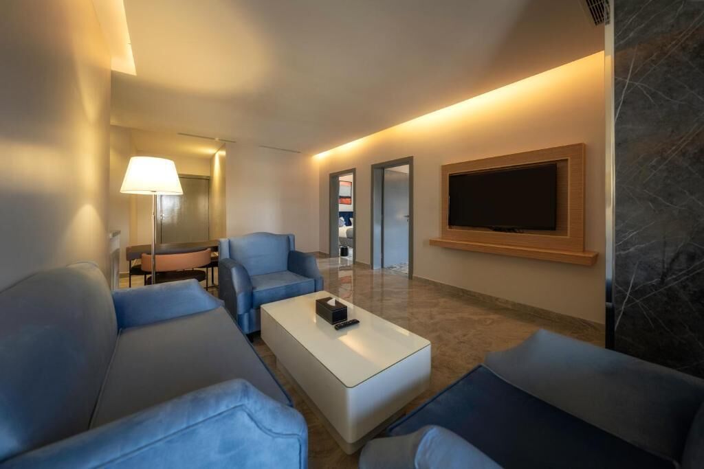 Una habitación de hotel moderna con sofá, una mesa de centro, un televisor de pantalla plana montado en la pared y una lámpara.