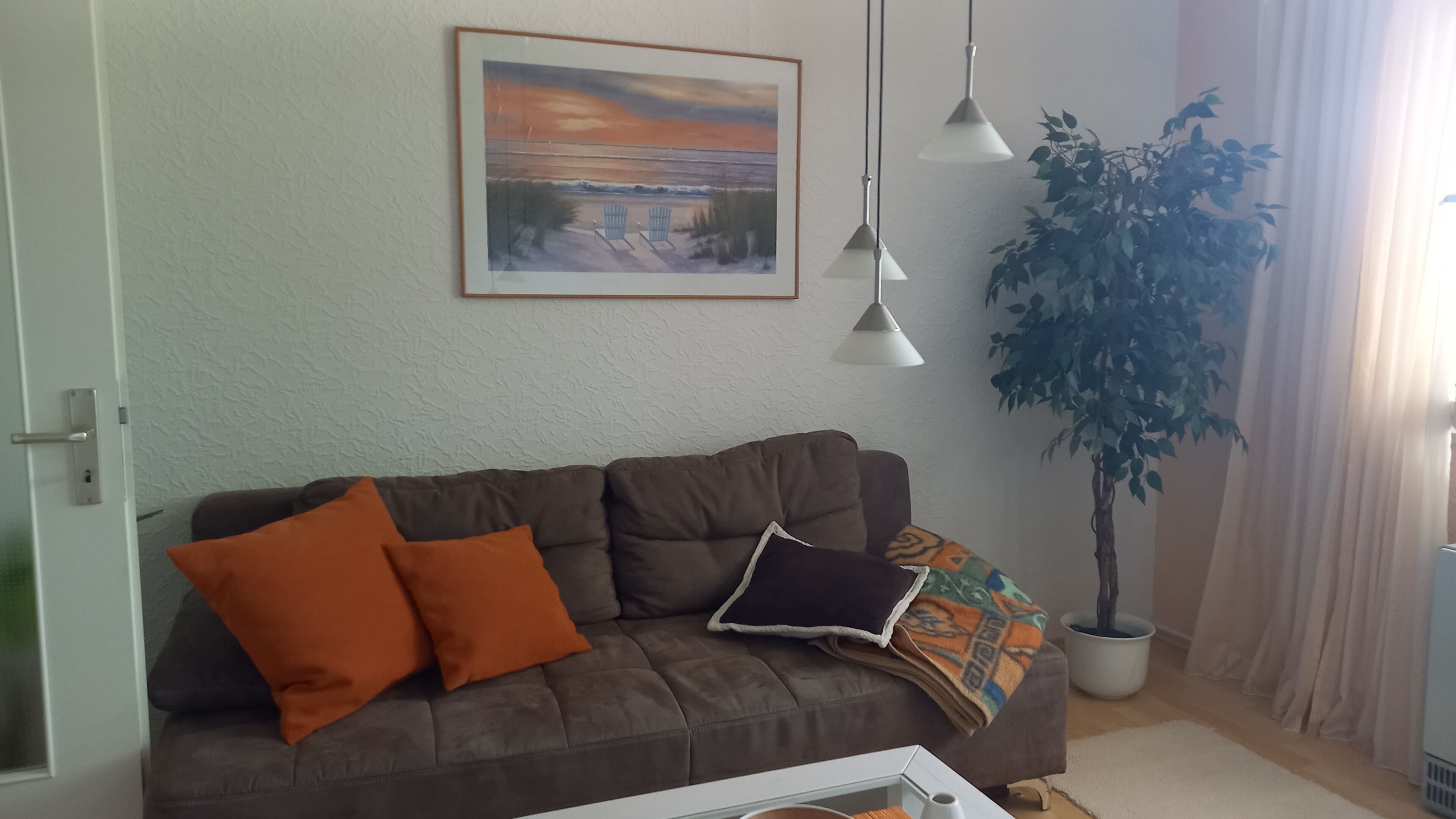 Ein Wohnzimmer mit einem braunen Sofa, zwei orangenen Kissen, einem gerahmten Bild, einer Topfpflanze und Pendelleuchten.