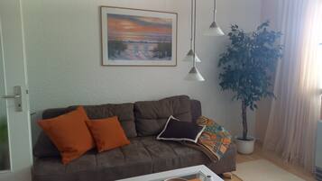 Ein Wohnzimmer mit einem braunen Sofa, zwei orangenen Kissen, einem gerahmten Bild, einer Topfpflanze und Pendelleuchten.