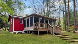 A cabin with a red exterior, a wooden deck, and a staircase leading to a covered porch. - Grace on the Lake-Little Red Cabin-Perch Lake, MI (South Boardman)
