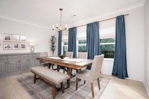 Un comedor con una mesa de madera, sillas, un banco, una araña, cortinas y cuadros en la pared.