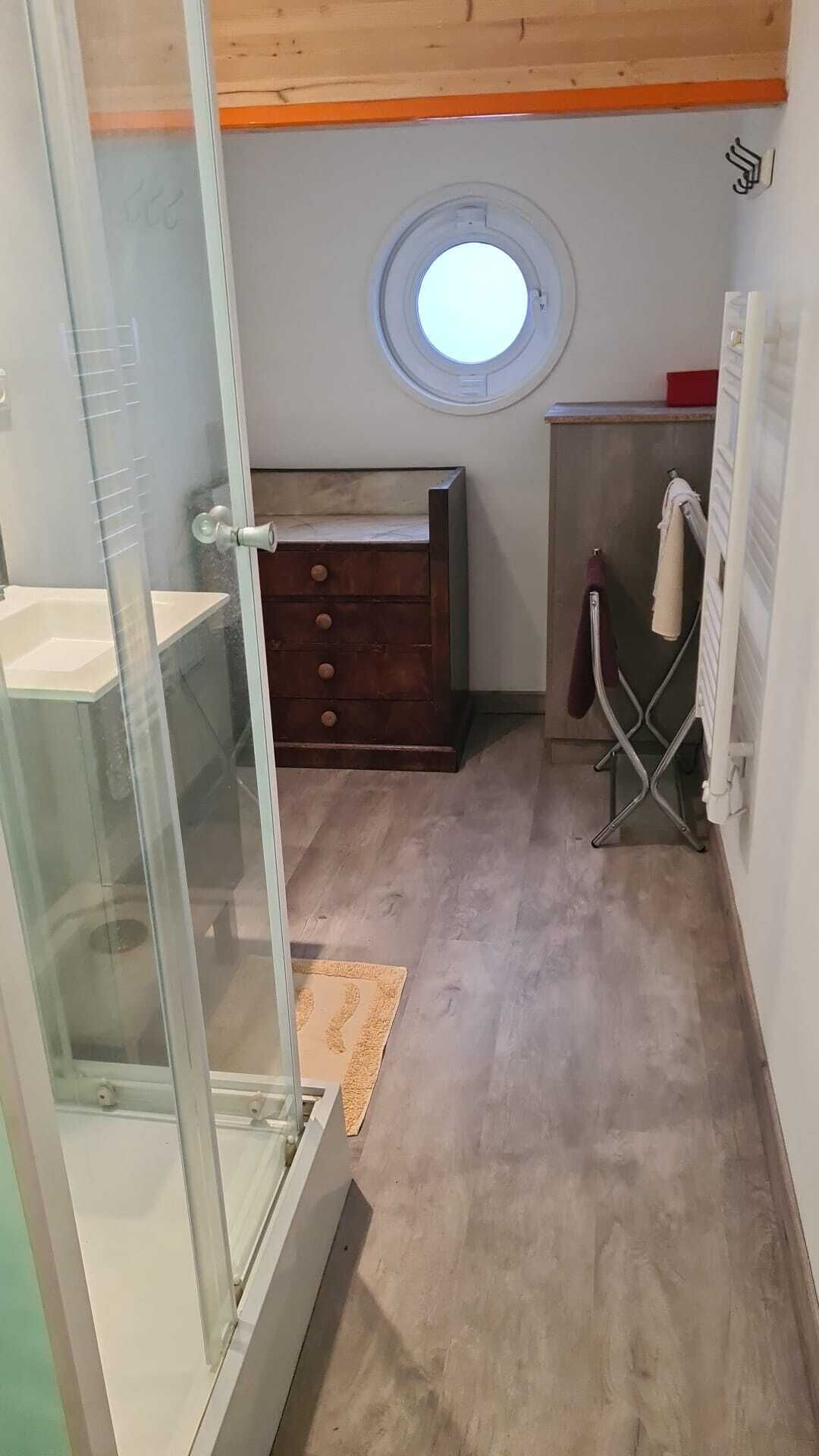 Un baño con cabina de ducha de vidrio, cómoda de madera, tragaluz circular y un perchero para toallas.