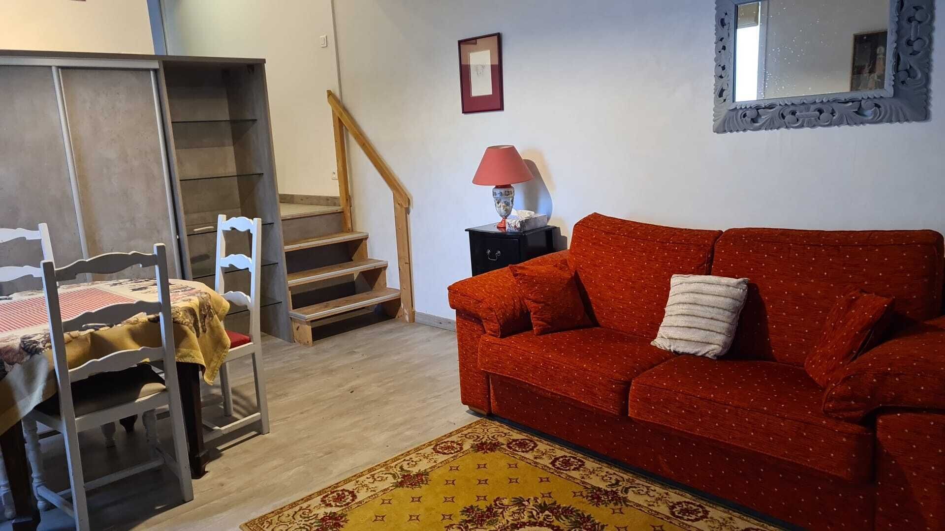 Una habitación con escalera de madera, un sofá rojo, una pequeña mesa con lámpara y una alfombra estampada.