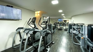 Un gimnasio con diversas máquinas de ejercicio, un televisor montado en la pared y un interior bien iluminado.