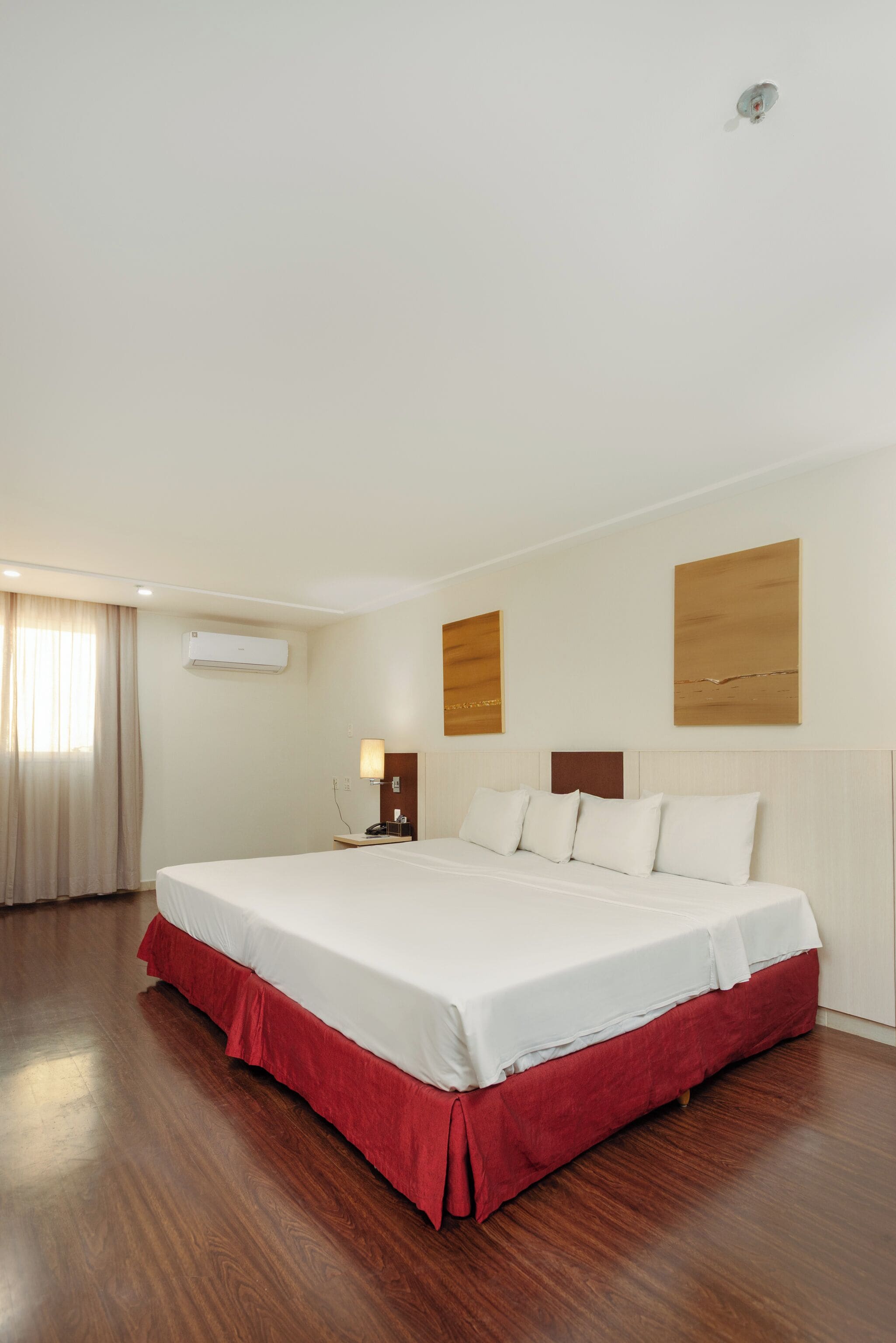 A hotel room with a large bed, a bedside table, an air conditioning unit, and a wall-mounted artwork.