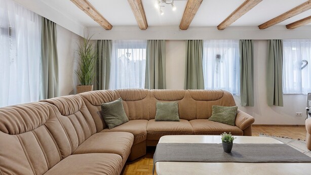 Ein Wohnzimmer mit einem Ecksofa, einem Couchtisch, Holzbalken an der Decke und einer Zimmerpflanze.