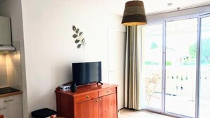 A compact living space with a kitchenette, a dining area, a television on a wooden cabinet, and a large sliding glass door leading to an outdoor area. - Appt T4 Residence Le Cordouan 3* pool side (Soulac-sur-Mer)