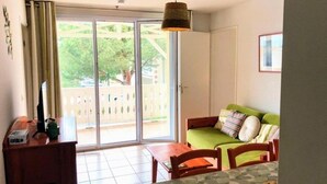 A room with a sliding glass door leading to a balcony, a green sofa with patterned cushions, a wooden table, and a hanging lamp. - Appt T4 Residence Le Cordouan 3* pool side (Soulac-sur-Mer)