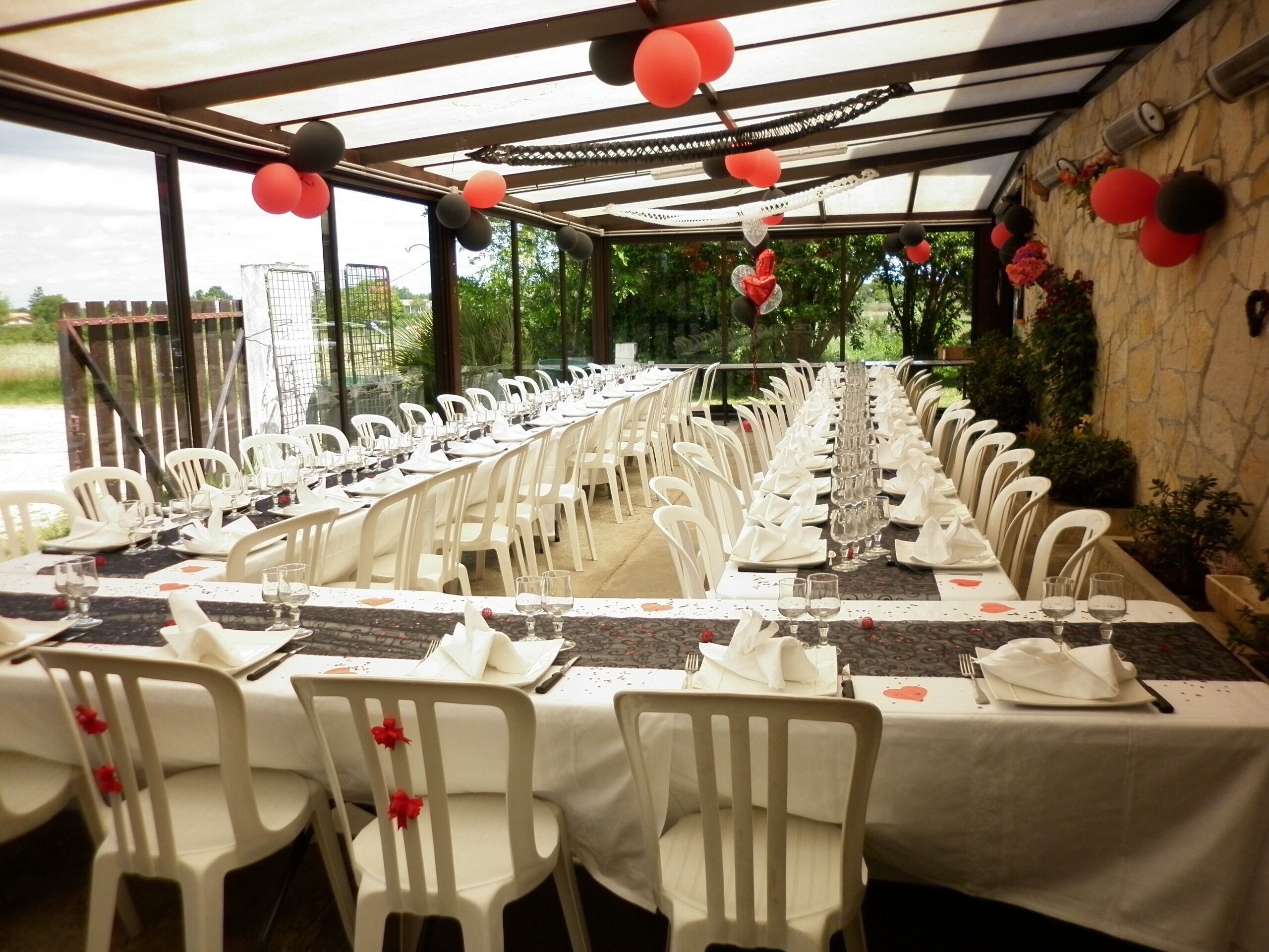 A long dining table set with white chairs, glassware, and plates, under a covered area with a stone wall and greenery visible outside.