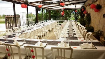 A long dining table set with white chairs, glassware, and plates, under a covered area with a stone wall and greenery visible outside.