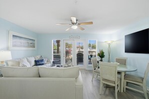 A living room with a sofa, a dining table with chairs, a ceiling fan, and a TV mounted on the wall. - Grand Caribbean East 107 (Pensacola)