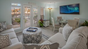 Ein Wohnzimmer mit Deckenventilator, einem Esstisch mit Stühlen, einem Gemälde der Aurora Borealis und Blick durch Glastüren auf einen Außenbereich mit dem Schild 'BEACH HOUSE'.