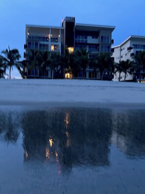 Een meerhoog hotel met een plat dak, palmbomen en een strand op de voorgrond.
