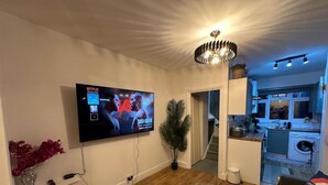 Een woonkamer met een flatscreen televisie aan de muur, een potplant, een trap die naar een keuken leidt, en een bank met een kussen.