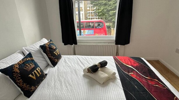 Een bed met wit beddengoed, twee kussens met 'VIP' geborduurd, een rode deken, en uitzicht vanuit het raam op een rode bus.