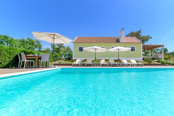 Una piscina con sillones de descanso y sombrillas, un edificio de tejado con baldosas y follaje al fondo.