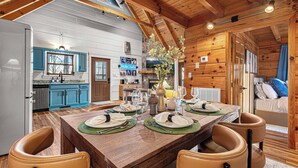 A wooden cabin interior with a dining table set for four, a kitchen with blue cabinets, and a cozy sleeping area. - Romantic Mtn Spa Retreat - Views • Hot Tub • Sauna • Fire Pit  • Cold Plunge (Gatlinburg)