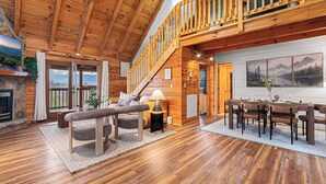 A spacious log cabin interior with a dining area, a fireplace, and a staircase leading to a loft. - Romantic Mtn Spa Retreat - Views • Hot Tub • Sauna • Fire Pit  • Cold Plunge (Gatlinburg)