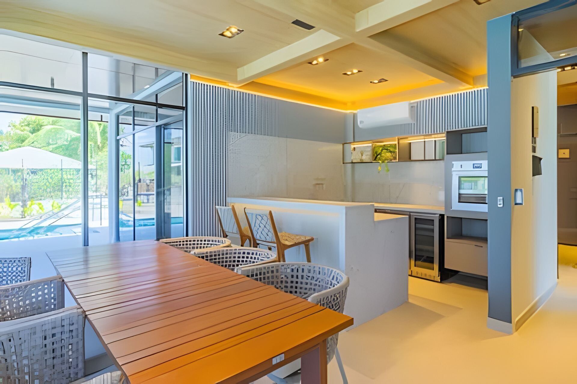 A modern kitchen with a central island, a dining area with a wooden table and wicker chairs, and a view of a swimming pool through large windows.