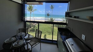 Un balcon avec vue sur la plage et l’océan, une table à manger avec assiettes et verres, et un coin cuisine équipé d’un barbecue.