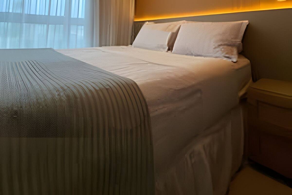 Una habitación de hotel moderna con una cama grande, una mesita de noche y un ventanal con cortinas translúcidas.