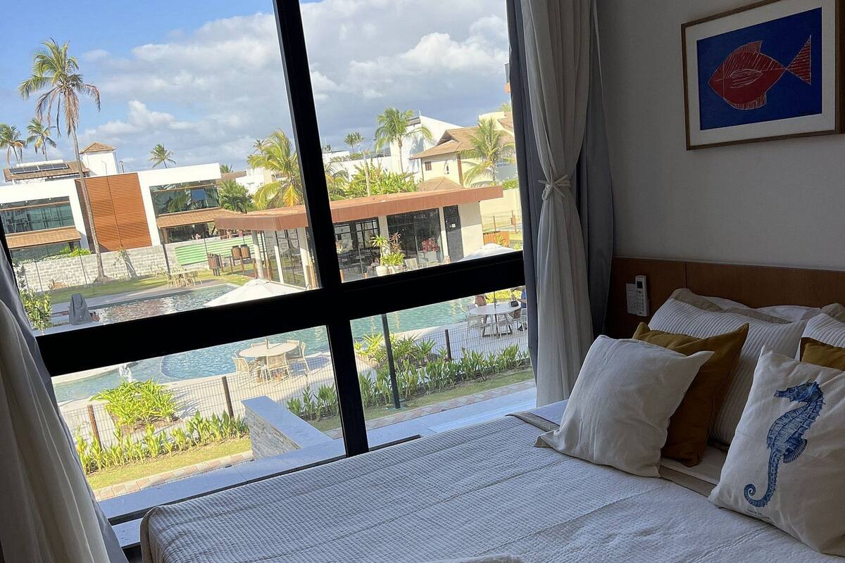 Habitación de hotel con un ventanal grande, cama con ropa de cama blanca y vista a una piscina y edificios.