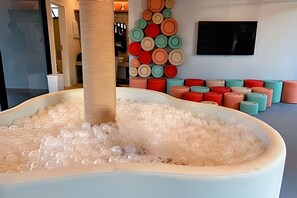 Una piscina grande, blanca y ovalada llena de agua y burbujas, rodeada de taburetes cilíndricos de colores.