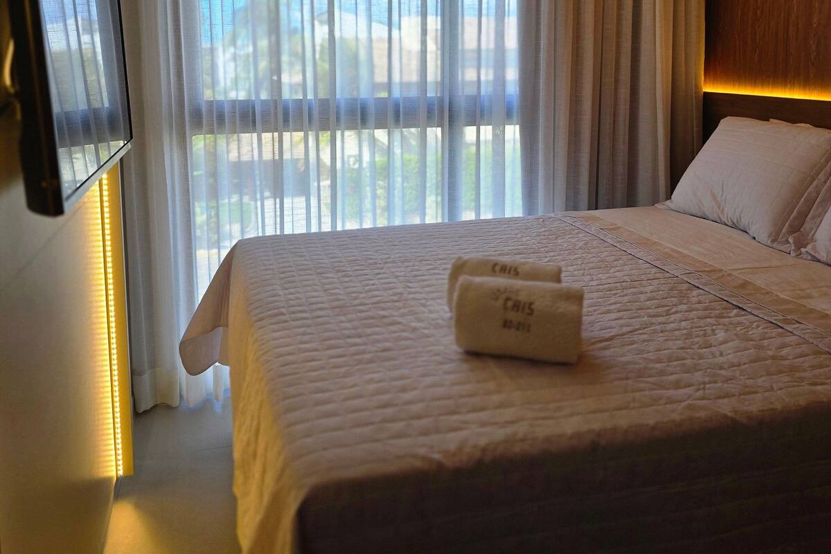Habitación de hotel con cama, televisor montado en la pared y un ventanal con cortinas.