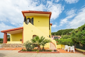 A yellow building with a red roof, a staircase, and a patio area with chairs.