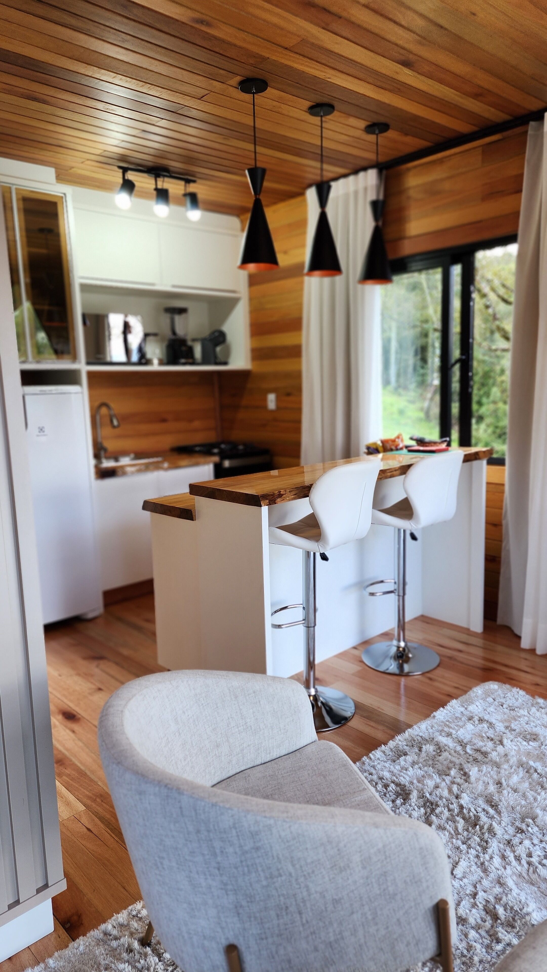 A modern kitchen with wooden ceiling, pendant lights, a dining area with white chairs, and a view through a window.