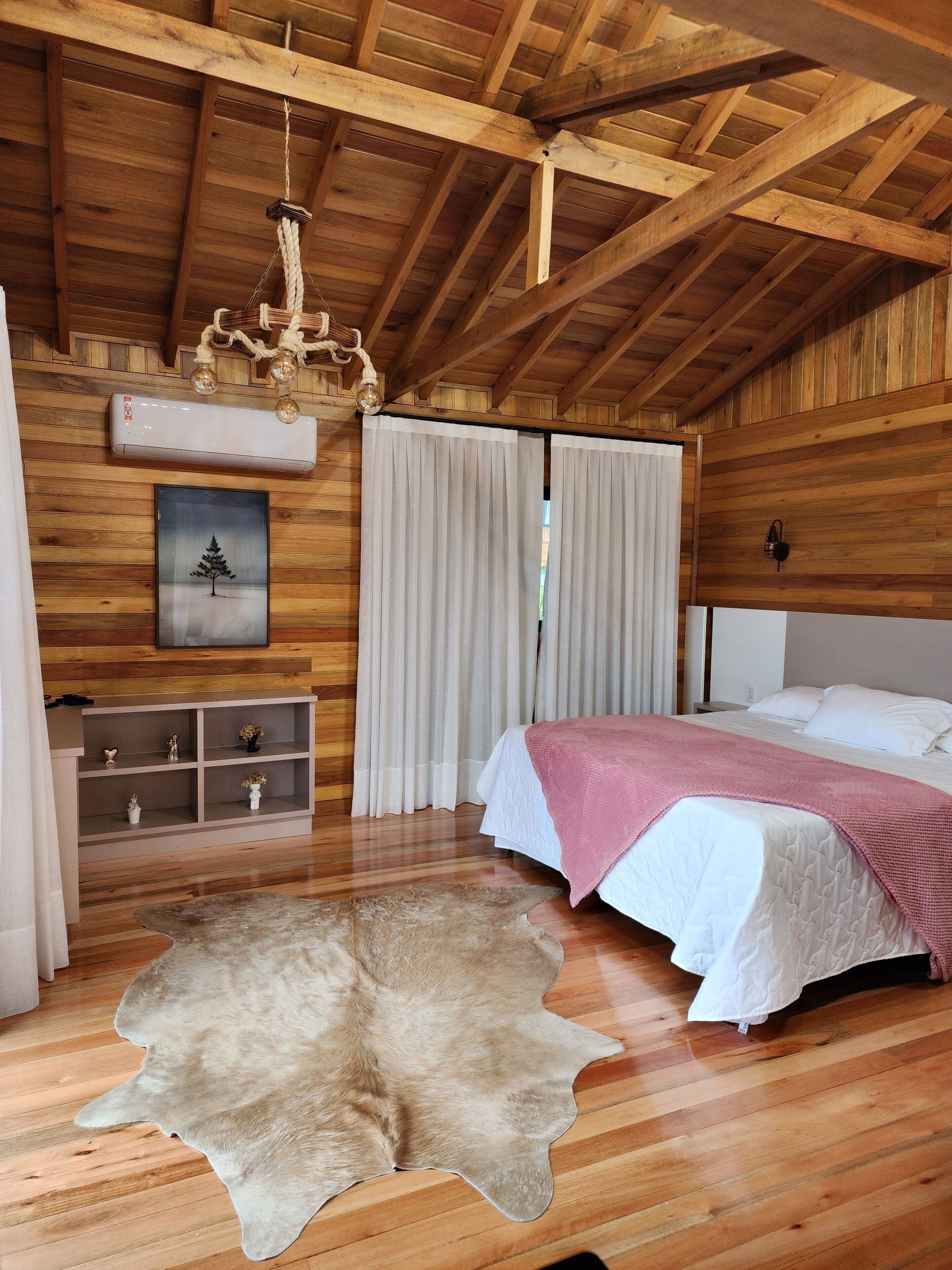 A wooden interior with a bed, curtains, a chandelier, a mounted air conditioner, and a mounted picture.