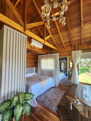 A wooden cabin bedroom with two beds, a chandelier, an air conditioning unit, and a view of the outdoors. - Pousada Pinha Pinhão (Urubici)