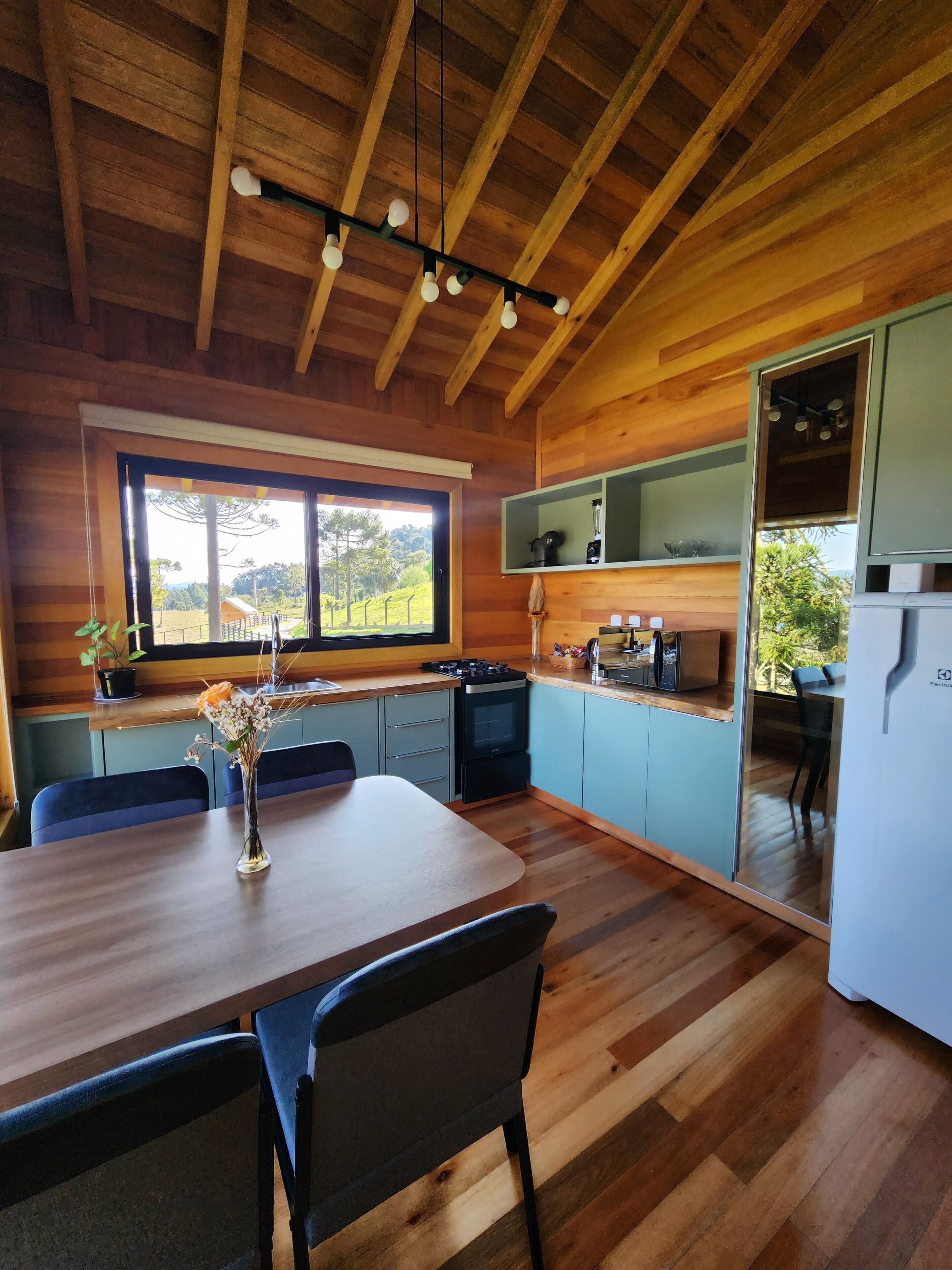 Un intérieur en bois comprenant une table à manger, des chaises, un coin cuisine équipé d’appareils électroménagers et une fenêtre donnant sur un paysage verdoyant.