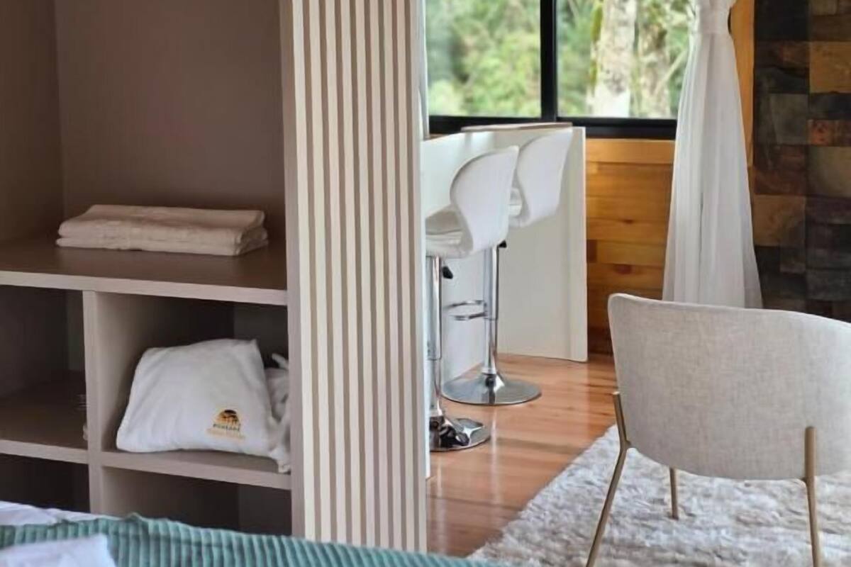 A room with a bed, a chair, a desk, a shelf with towels, and a window with curtains.