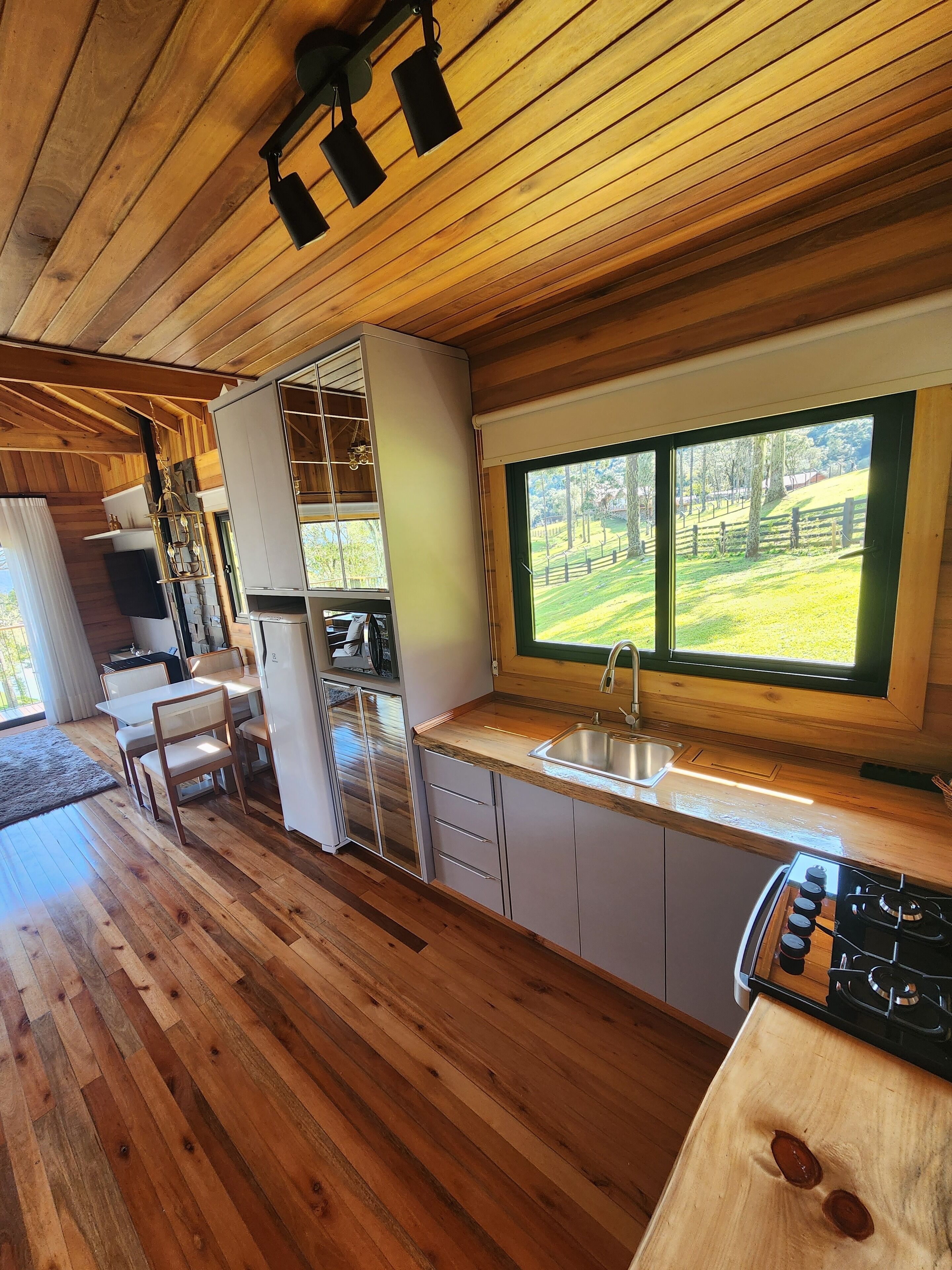 A wooden interior with a kitchen, dining area, and a large window overlooking a green landscape.