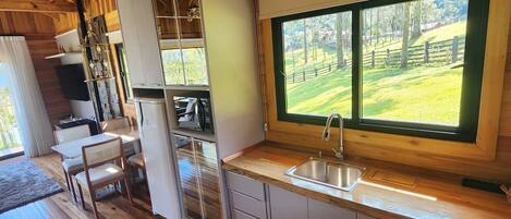 A wooden interior with a kitchen, dining area, and a large window overlooking a green landscape.