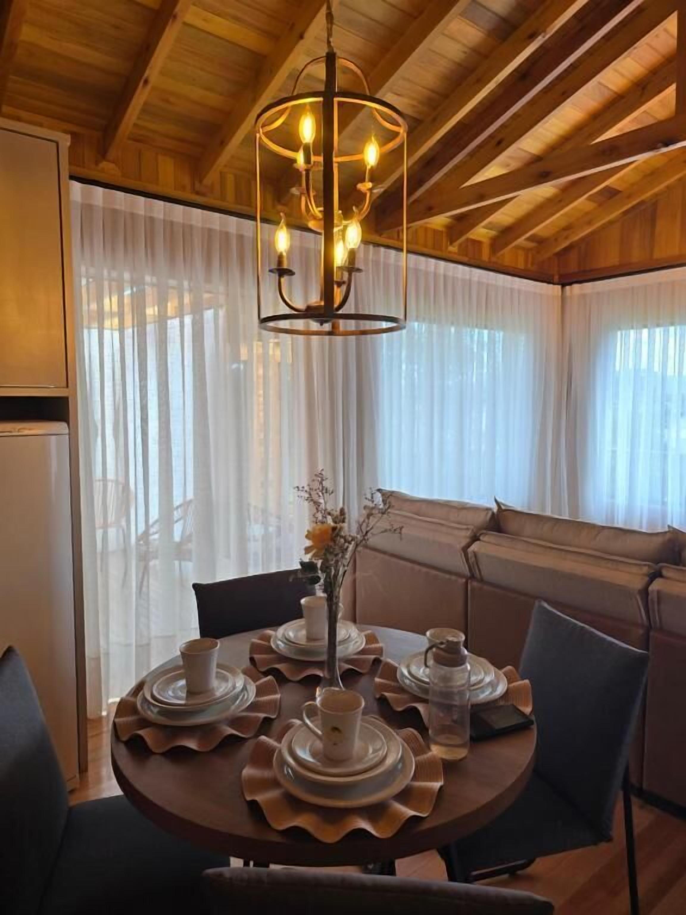 Un intérieur en bois avec une table de salle à manger ronde, disposée pour accueillir quatre personnes, un lustre et un canapé.