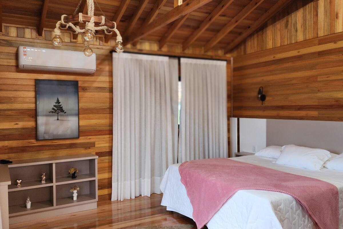 A wooden-paneled room with a bed, a chandelier, a mounted air conditioner, a shelf with decorative items, and a fur rug on the floor.