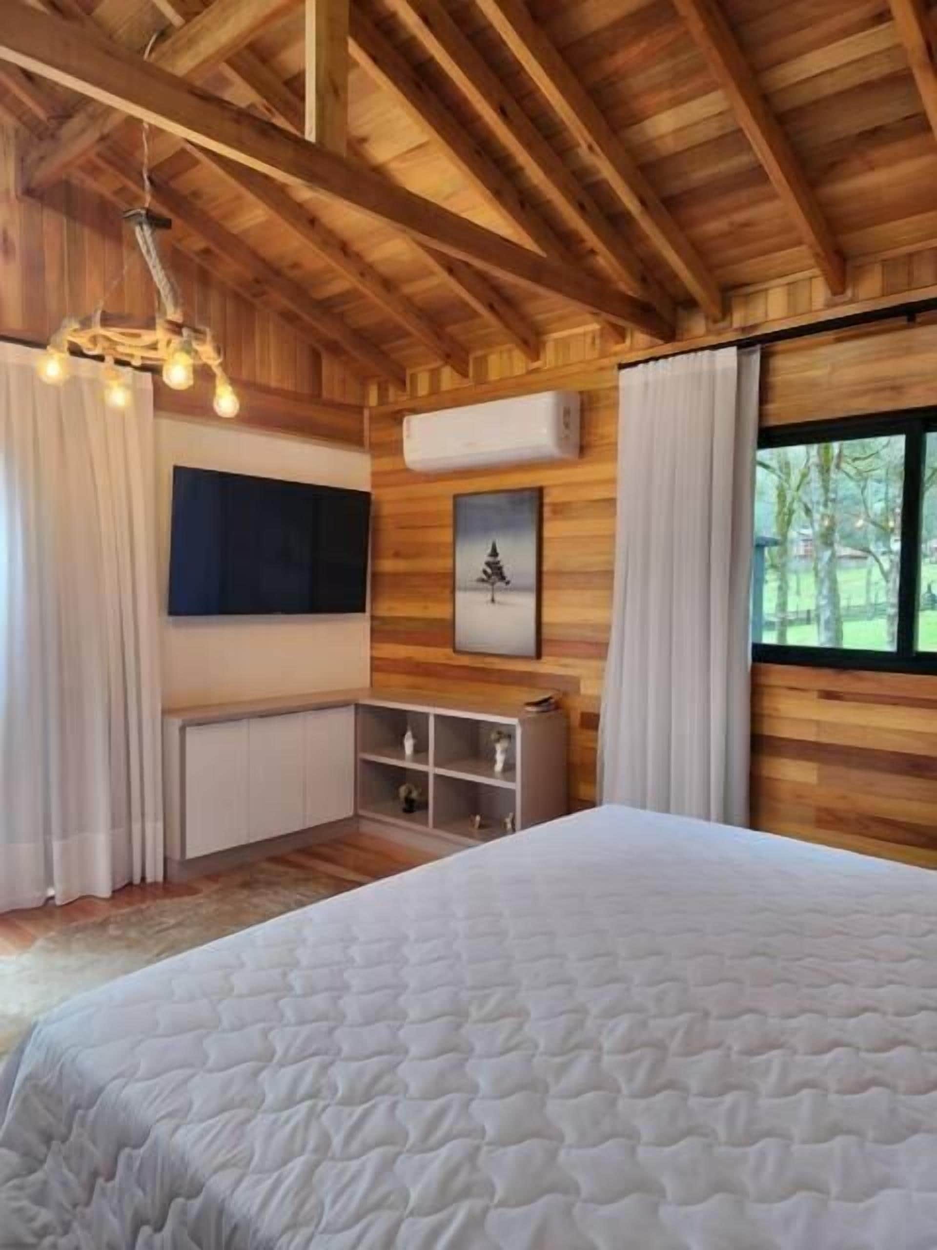 Une chambre dans une cabane en bois, avec un lit, un téléviseur fixé au mur, une étagère avec des objets décoratifs et une fenêtre avec des rideaux.