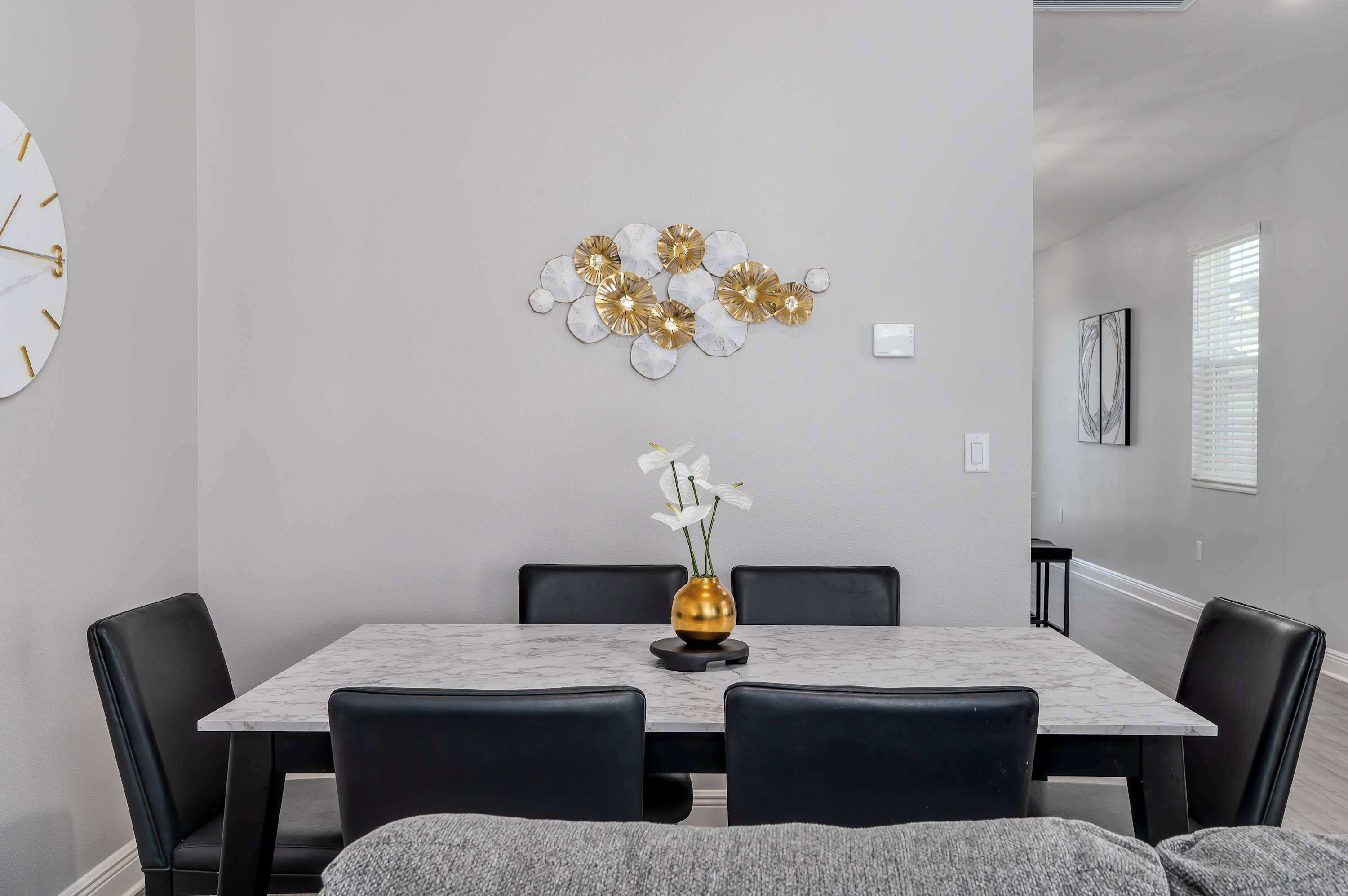 A dining area with a marble table, black chairs, a gold vase with white flowers, and a wall-mounted abstract art piece.