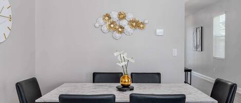 A dining area with a marble table, black chairs, a gold vase with white flowers, and a wall-mounted abstract art piece.