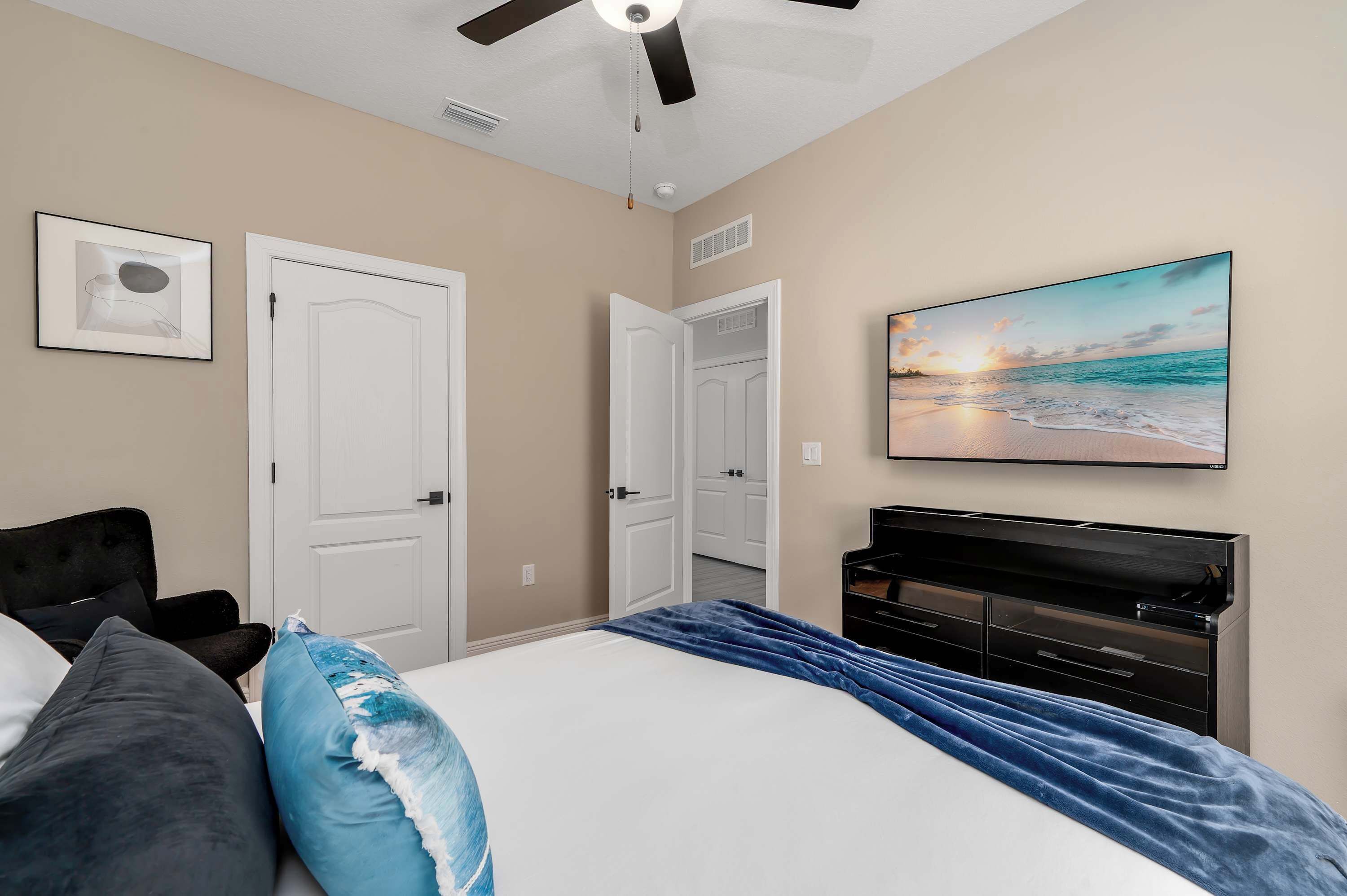 A bedroom with a bed, a ceiling fan, a painting of a beach scene, and a black dresser.
