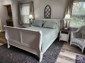 A bedroom with a bed, two bedside tables, a chair, a window with curtains, and a wall-mounted decorative piece.