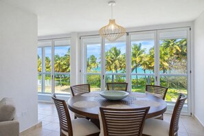 A dining area with a round table, chairs, and a bowl centerpiece. Large windows offer a view of palm trees and the ocean. - Beachfront Penthouse with Private Terrace, Ocean & El Yunque Views – Casa Bacci (Río Grande)