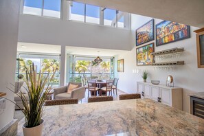 A spacious kitchen with a large island, dining area with chairs, and a view of palm trees through large windows. - Beachfront Penthouse with Private Terrace, Ocean & El Yunque Views – Casa Bacci (Río Grande)